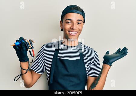 Giovane bell'artista di tatuaggio dell'uomo afroamericano che indossa l'uniforme professionale e guanti che tengono la macchina tatooer sorridendo allegro presentazione e poi Foto Stock