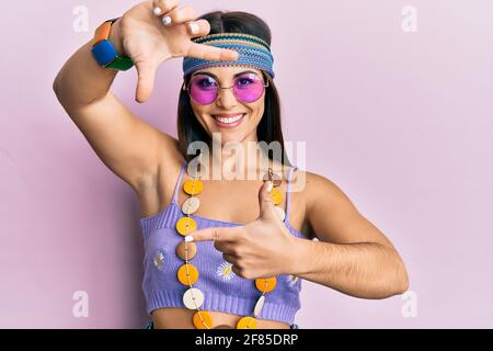 Giovane donna bruna con stile bohémien e hippie sorridente che fa cornice con mani e dita con volto felice. Creatività e concetto di fotografia Foto Stock