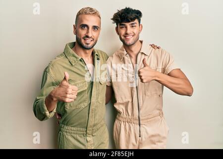 Coppia omosessuale gay in piedi insieme indossando jumpsuit casual facendo felice pollici su gesto con la mano. Espressione di approvazione guardando la macchina fotografica sh Foto Stock