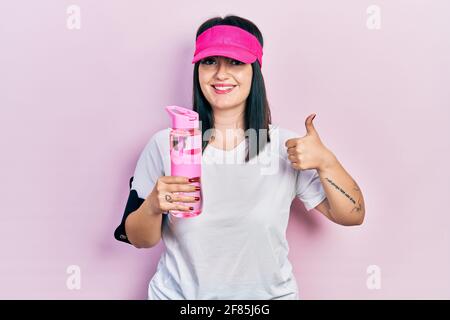 Giovane donna ispanica che indossa sportswear bere bottiglia d'acqua sorridente felice e positivo, pollice su facendo eccellente e segno di approvazione Foto Stock