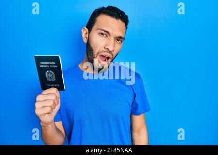 Uomo ispanico con barba che tiene il passaporto italia che guarda sonnolento e stanco, esausto per stanchezza e sbornia, occhi pigri al mattino. Foto Stock