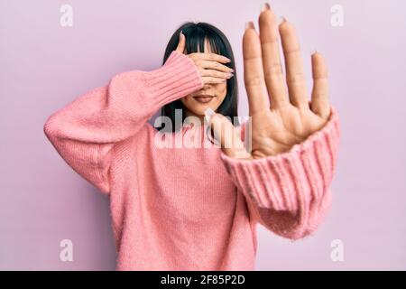 Giovane donna bruna con pangs che indossa un maglione invernale casual che copre gli occhi con le mani e fa il gesto di stop con espressione triste e paura. Imbarazzo Foto Stock