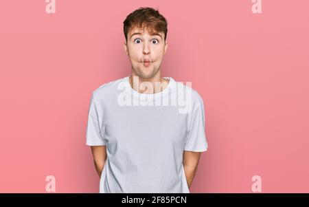 Giovane uomo caucasico che indossa una t-shirt bianca casual che fa viso di pesce con labbra, gesti pazzi e comici. Espressione divertente. Foto Stock