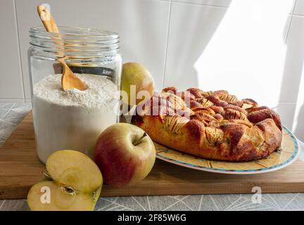 Una torta di mele in un piatto su una tavola di legno accanto ad un vaso di farina e alcune mele Foto Stock