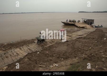 Kampong Cham, Cambogia. Un traghetto che aspetta i passeggeri a bordo, mentre le persone che trasportano merci che si spostano sul sentiero in un porto di attraversamento del fiume Mekong, con Koh Pen Island può essere visto in parte sullo sfondo. Il fiume Mekong, che scorre per quasi 4,763 km in sei paesi, è un canale navigabile importante sia per il trasporto di merci che di passeggeri, secondo la Mekong River Commission (MRC) nella loro strategia di sviluppo del bacino del 2021-2030 e nel piano strategico del 2021-2025. Foto Stock