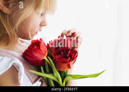 Piccola ragazza sorridente che tiene bouquet di fiori rossi di tulipano. Concetto di biglietto d'auguri per Pasqua, Festa della Madre, Giornata Internazionale della Donna, San Valentin Foto Stock