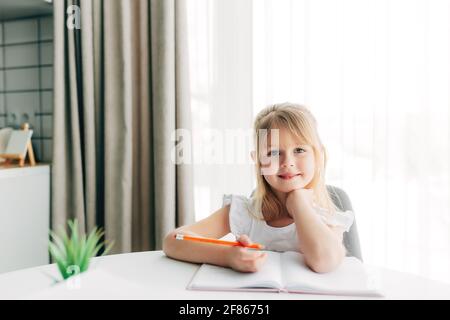 Una piccola ragazza sorridente si siede al tavolo e scrive in un taccuino bianco. Concetto di istruzione. Scuola domestica. Compiti. Faccia sorridente. Foto di alta qualità Foto Stock