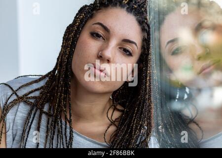 Giovane donna con dreadlock e piercing al naso che pende contro un finestra con riflesso sul vetro che guarda attentamente fotocamera in primo piano Foto Stock