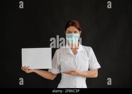 Un giovane medico in una maschera medica tiene un poster bianco su uno sfondo nero. Infermiera femminile in uniforme che punta ad un cartellone vuoto. Foto Stock