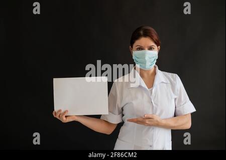 Un giovane medico in una maschera medica tiene un poster bianco su uno sfondo nero. Infermiera femminile in uniforme che punta ad un cartellone vuoto. Foto Stock