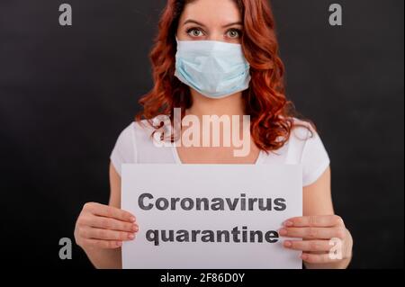 La donna spaventata in una maschera medica contiene un poster. Ragazza dai capelli rossi su sfondo nero in un respiratore di quarantena antimicrobico. Coronavirus COVID-19 Foto Stock