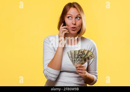 Ritratto di una donna caucasica sorprende un fan di dollari e parla al telefono. Sfondo giallo. Il concetto di sconti e vendite. Foto Stock