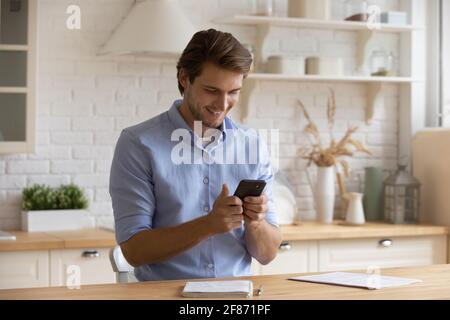 Uomo sorridente che tiene uno smartphone, guarda lo schermo, seduto in cucina Foto Stock