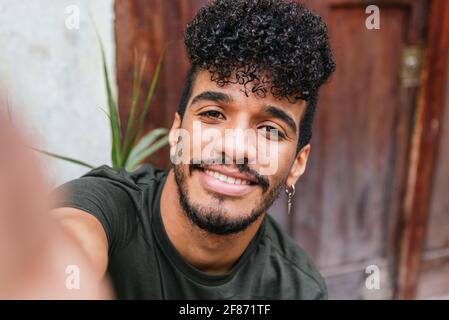 Autoritratto di giovane attraente latino con il suo cellulare. Indossa una t-shirt verde scuro e sorride alla telecamera. Concetto di spontaneità e gioia Foto Stock