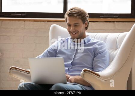 Primo piano uomo sorridente con il computer portatile, seduto in una comoda poltrona Foto Stock