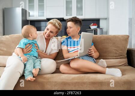 Madre sorridente con due figli di età diversa che giocano sul divano e usano un computer portatile moderno. Stile di vita domestico e tempo di famiglia. Foto Stock