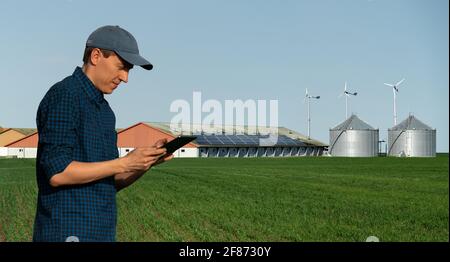 Agricoltore con computer tablet sullo sfondo di una moderna fattoria casearia che utilizza energia rinnovabile, pannelli solari e turbine eoliche Foto Stock