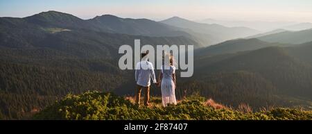 Vista posteriore di una coppia amorevole che si erge su una collina erbosa e tiene le mani mentre si guarda le belle montagne. Giovane uomo che tiene la mano di donna affascinante in abito bianco e ammirando la bellezza della natura. Foto Stock