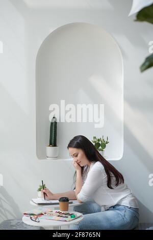 Bella giovane donna sorridente che beve caffè e disegnando ritratto con matite in un libro di schizzo Foto Stock