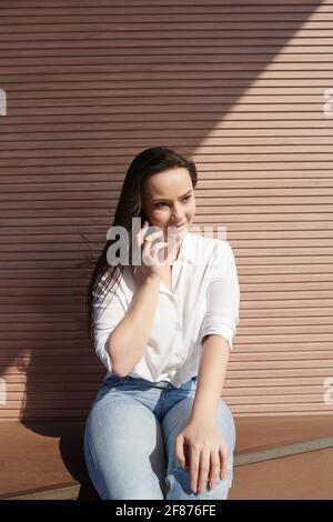 Ritratto di giovane donna attraente seduta all'aperto e che parla sopra telefona con gli amici che parlano di gossip Foto Stock