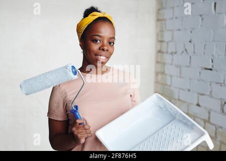Giovane donna africana allegra con lattrollatore in piedi in un angolo contro due pareti e guardarti con sorriso durante ristrutturazione della casa Foto Stock