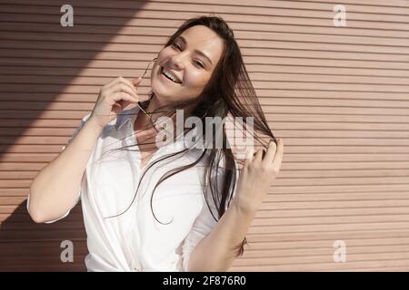 Ritratto di bella bruna giovane sorridente in piedi al muro della casa sul dat sole e guardando la macchina fotografica Foto Stock