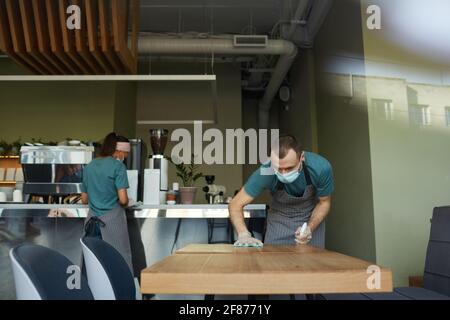 Ritratto di un giovane uomo che indossa una maschera mentre pulisce i tavoli con un igienizzatore al bar o al bar, covid misure di sicurezza, copy space Foto Stock