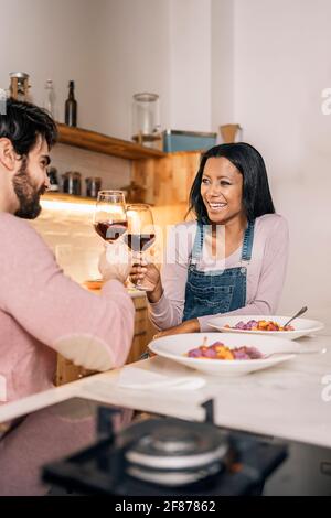Simpatica coppia multietnica che pranza insieme in cucina e beve un bicchiere di vino. Foto Stock