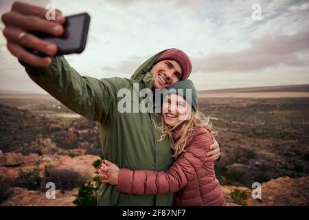 Giovane coppia allegra abbracciando durante il campeggio di montagna e prendendo selfie utilizzo dello smartphone su sfondo naturale Foto Stock