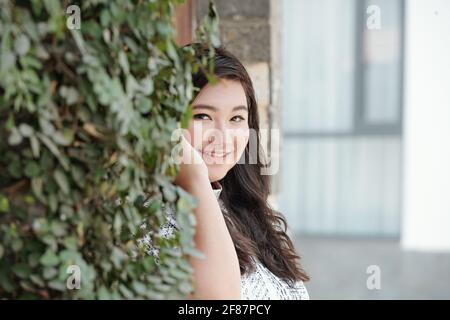 Ritratto di bella giovane donna sorridente in piedi al muro coperto con edera e sorridente alla macchina fotografica Foto Stock