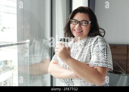 Ritratto di felice giovane donna in bicchieri in piedi presso l'hotel finestra della camera con una tazza di caffè da take-out in mano Foto Stock