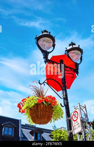 Petunias rosse in un cesto di fiori appeso ad un palo di luce stradale con un ombrello rosso decorativo. Decorazioni e ornamenti urbani. Foto Stock