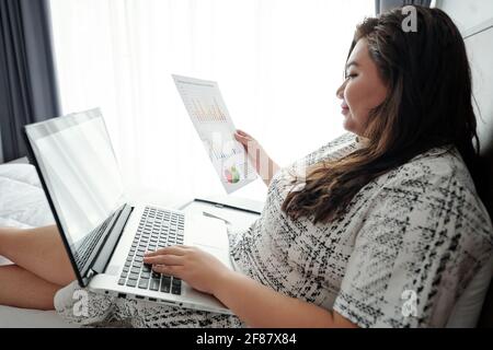 Una giovane donna d'affari sorridente seduta sul letto nella camera d'albergo e. analisi del documento con statistiche di vendita Foto Stock
