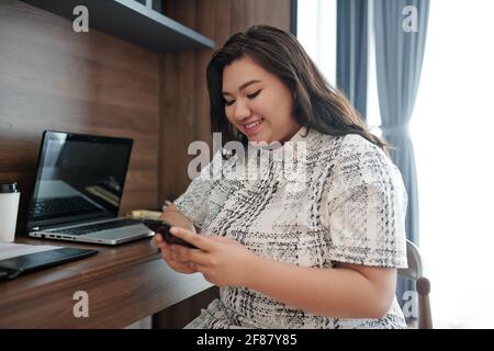 Imprenditore femminile piuttosto positivo seduto a tavola nella sua casa ufficio e la lettura di messaggi di testo da clienti felici Foto Stock