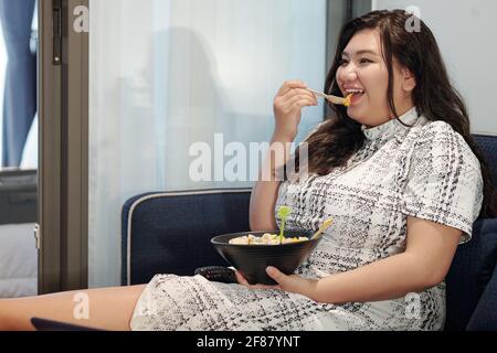Felice giovane donna che mangia deliziosa insalata di frutta quando watchong film comico in tv Foto Stock
