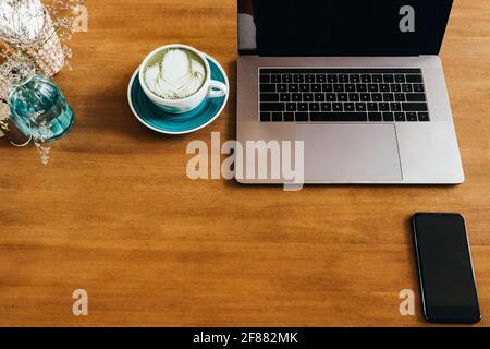Vista dall'alto del moderno computer portatile con telefono e dell'elegante tazza da caffè Matcha sul tavolo marrone in un bar leggero ed elegante. Spazio di copia. Foto Stock