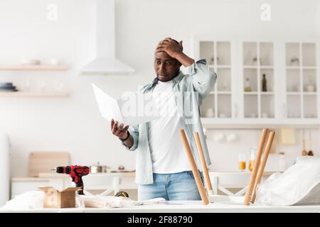 Difficoltà e problemi nell'assemblaggio di mobili nuovi. Confuso uomo afro-americano istruzione di lettura Foto Stock