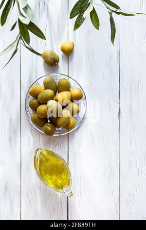 Olio d'oliva e ciotola di olive verdi su un tavolo di legno bianco. Moderna e minimal still life, ciotola in vetro con olio e olive, rametto di olive e spazio per fotocopie. Foto Stock