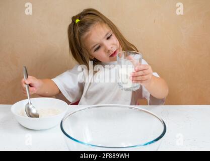 Una bambina vestita come cuoco impasta l'impasto. Cucina bambino stile di vita concetto. Il capretto ama, ha divertimento, studia e gioca nella cucina Foto Stock