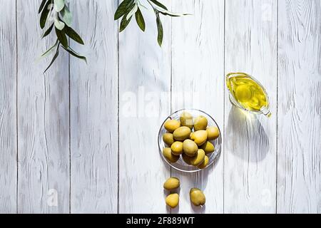 Olio d'oliva e ciotola di olive verdi su un tavolo di legno bianco. Moderna e minimal still life, ciotola in vetro con olio e olive, rametto di olive e spazio per fotocopie. Foto Stock