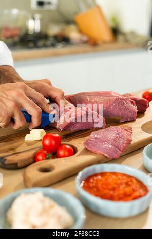 Un primo piano delle mani dello chef tagliando la carne a pezzi sottili con un coltello. Varie spezie e verdure possono essere viste intorno. Foto Stock