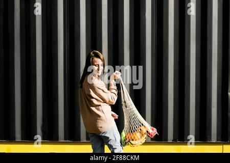 Colori alla moda senza sprechi. Allegra giovane donna sorridente e che tiene riutilizzabile borsa della spesa in rete con frutta e verdura. Foto Stock