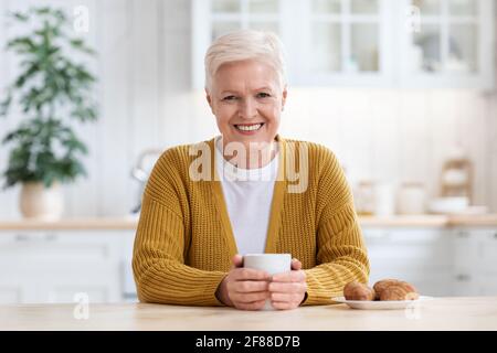 Signora anziana allegra che beve caffè con croissant Foto Stock