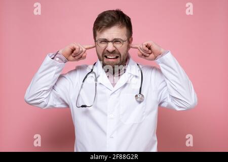 Medico con uno stetoscopio intorno al collo è stressato, è infastidito dal rumore, copre le orecchie con le dita e i denti di lacrime. Foto Stock