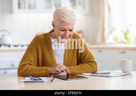 Donna anziana con capelli grigi positiva che usa il tablet digitale in cucina Foto Stock