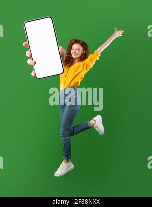 Offerta speciale, vendita online. Giovane donna spensierata che salta in aria, mostrando cellulare con spazio mockup su sfondo verde Foto Stock