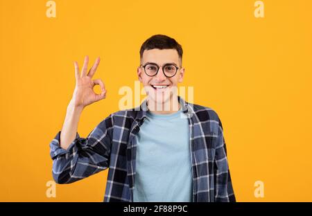 Ritratto di felice ragazzo millenario in occhiali mostrando un gesto OK su sfondo arancione studio Foto Stock