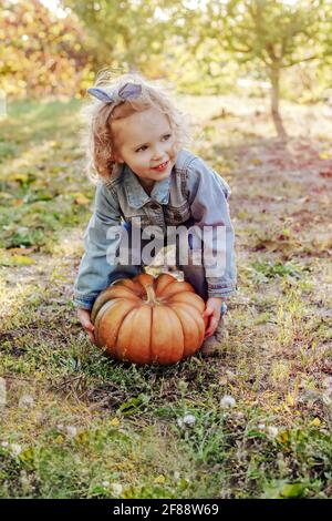 Ragazzina cercando di sollevare la zucca enorme all'aperto. Zucca di Halloween in via d'autunno in bambini mani di bambina bionda caucasica in denim suit in f Foto Stock