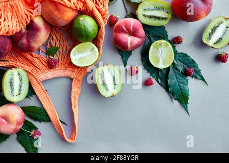 Stay piatto di eco-friendly mesh shopping bag con frutta lampone, pesca, kiwi, lime su sfondo grigio in luce solare, estate. Concetto di drogheria, poliziotto Foto Stock