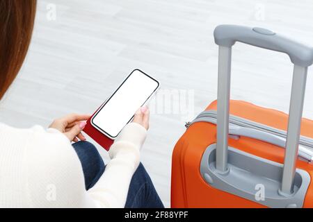 Vista posteriore di ragazza che tiene smartphone con bianco schermo mockup. Viaggiatore con telefono all'aeroporto con passaporto e valigia. Concetto di stile di vita Foto Stock